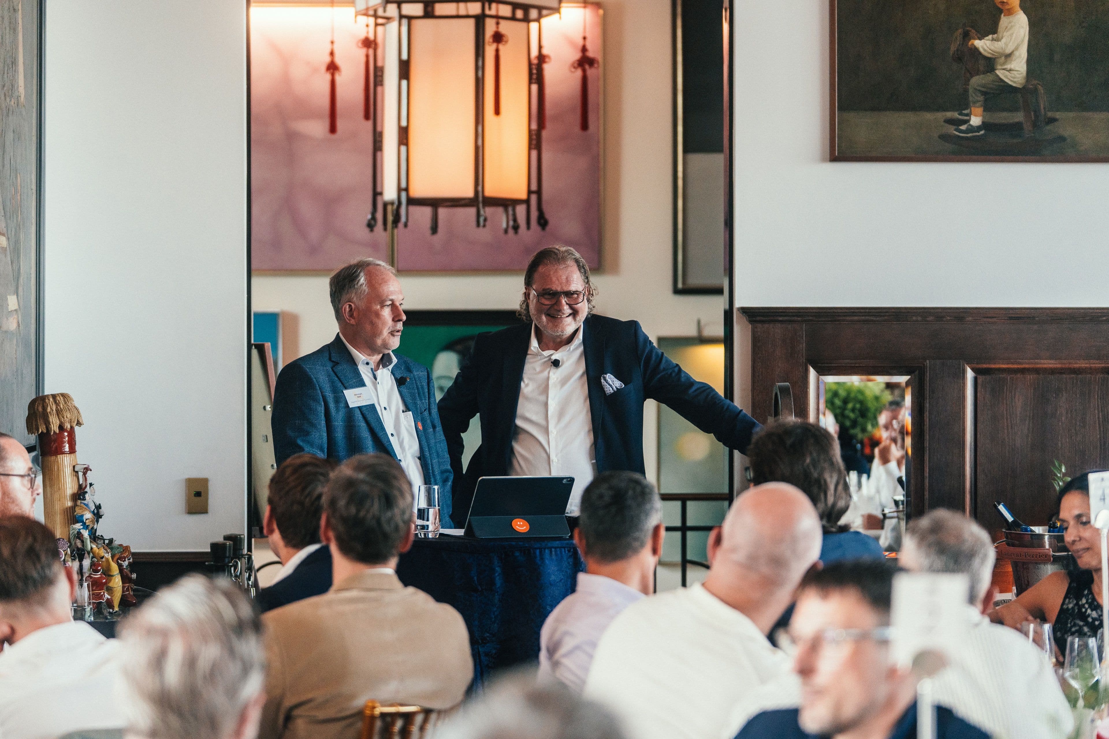 Zwei Redner sprechen vom Podium zu den sitzenden Gästen